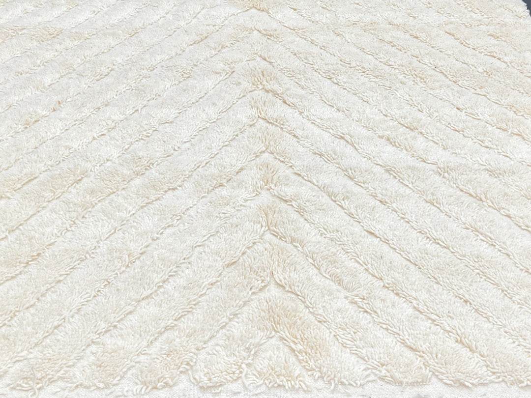 Beige textured rug with tassels on a black floor against a light gray brick wall.